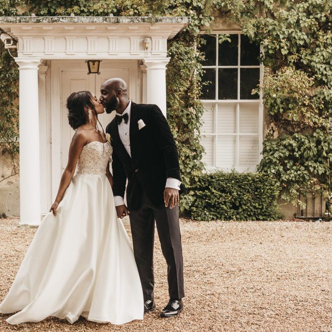 Black bride in a strapless Oleg Cassini wedding dress with beaded bodice kissing her groom in a velvet tuxedo and bow tie outside Northbrook Park 