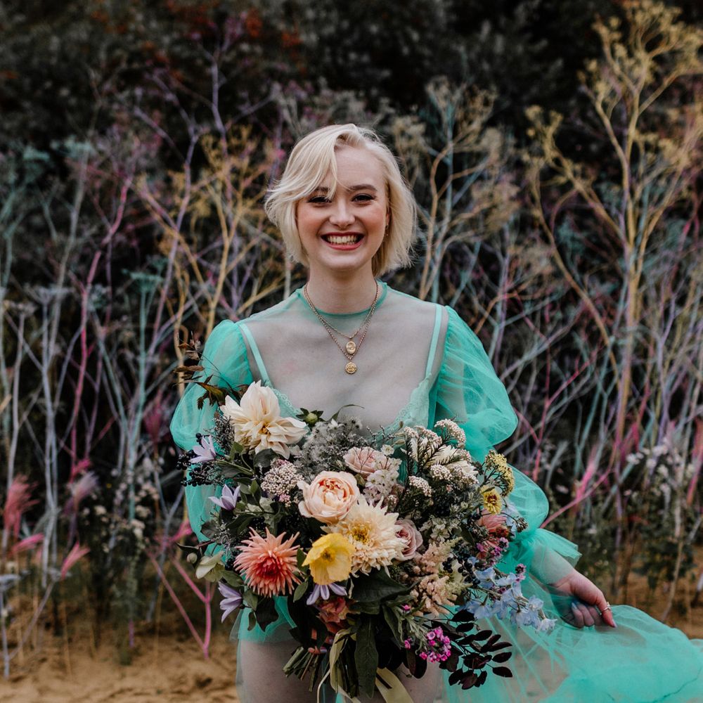 White blonde bride with bobbed hair in a sheer green pastel wedding dress holding a colourful wedding bouquet tied with ribbon