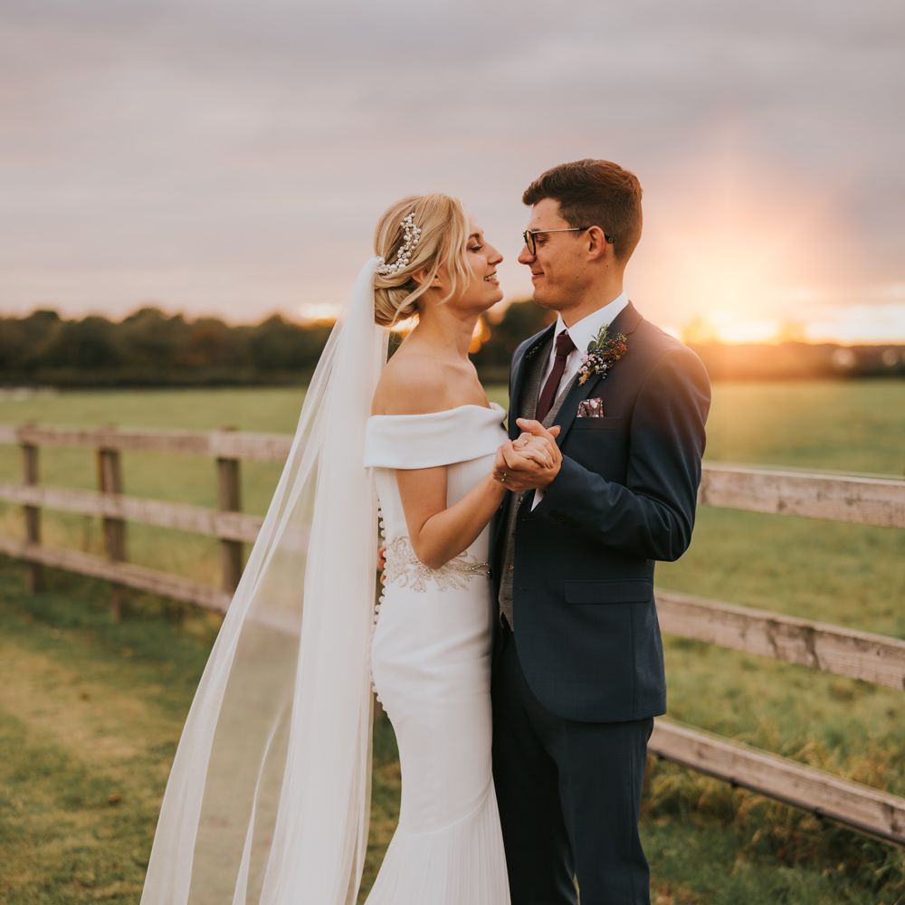 Bride & groom look lovingly at one another in the countryside as the sun sets in the background
