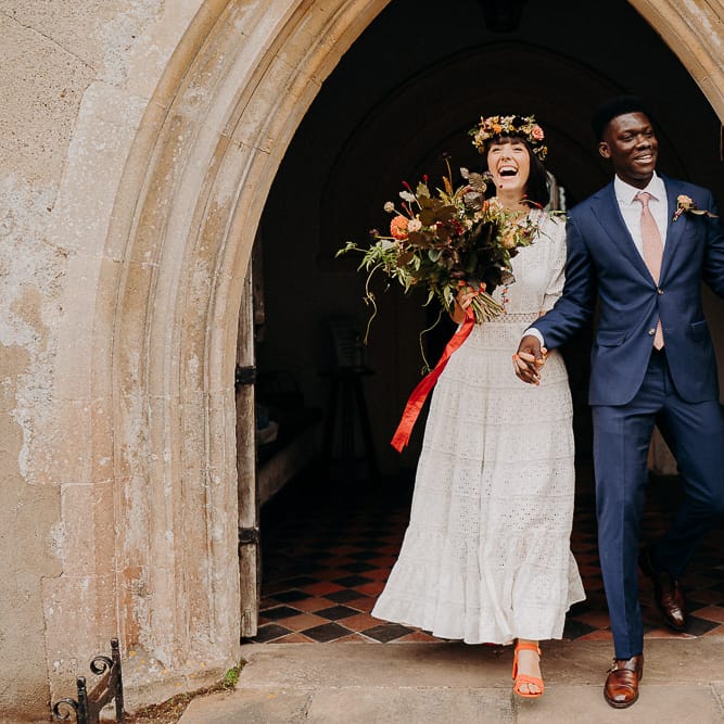 Bride in a Broderie Anglaise wedding dress and flower crown and groom in a navy suit exiting the church, just married 