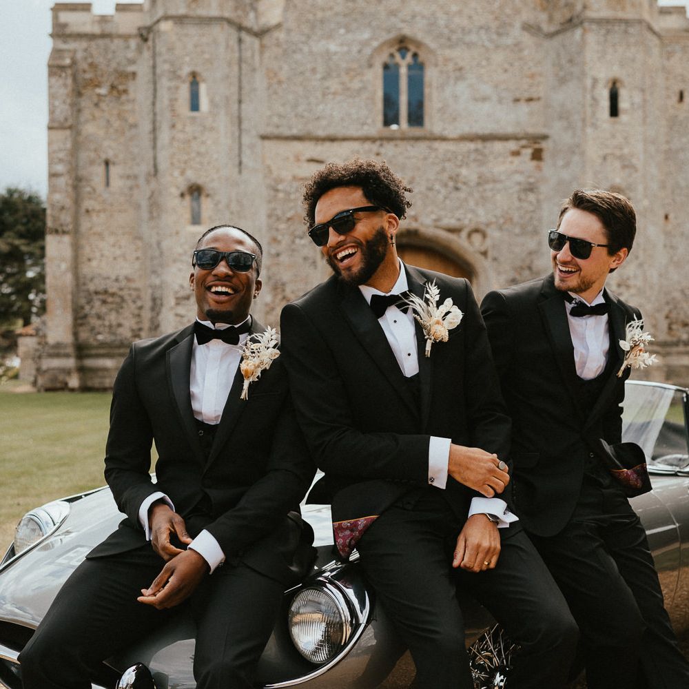 Happy groom and groomsmen in three piece black suits with dried flower buttonholes laughing whilst sitting on the bonnet of the classic wedding car 