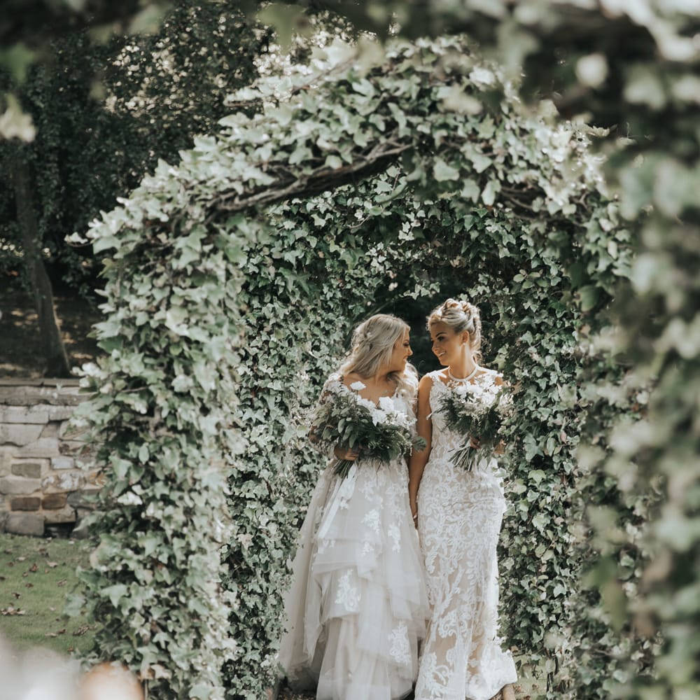 Two brides embrace under ivy covered arches at Crab & Lobster fairytale wedding.