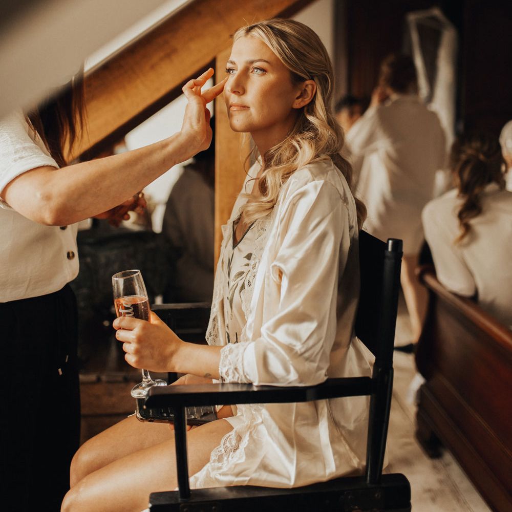 Bride with curled blonde hair in satin lace trimmed bridal dressing gown sits on chair holding a flute of sparkling wine as she has her make up done before wedding at Anran Devon
