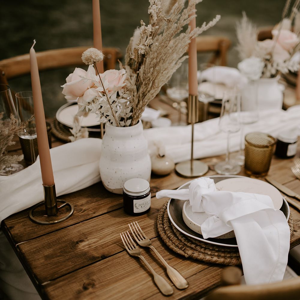 Boho wedding reception table decor with wicker place mat, linen napkin, gold cutlery and vases filled with neutral dried and fresh flowers 