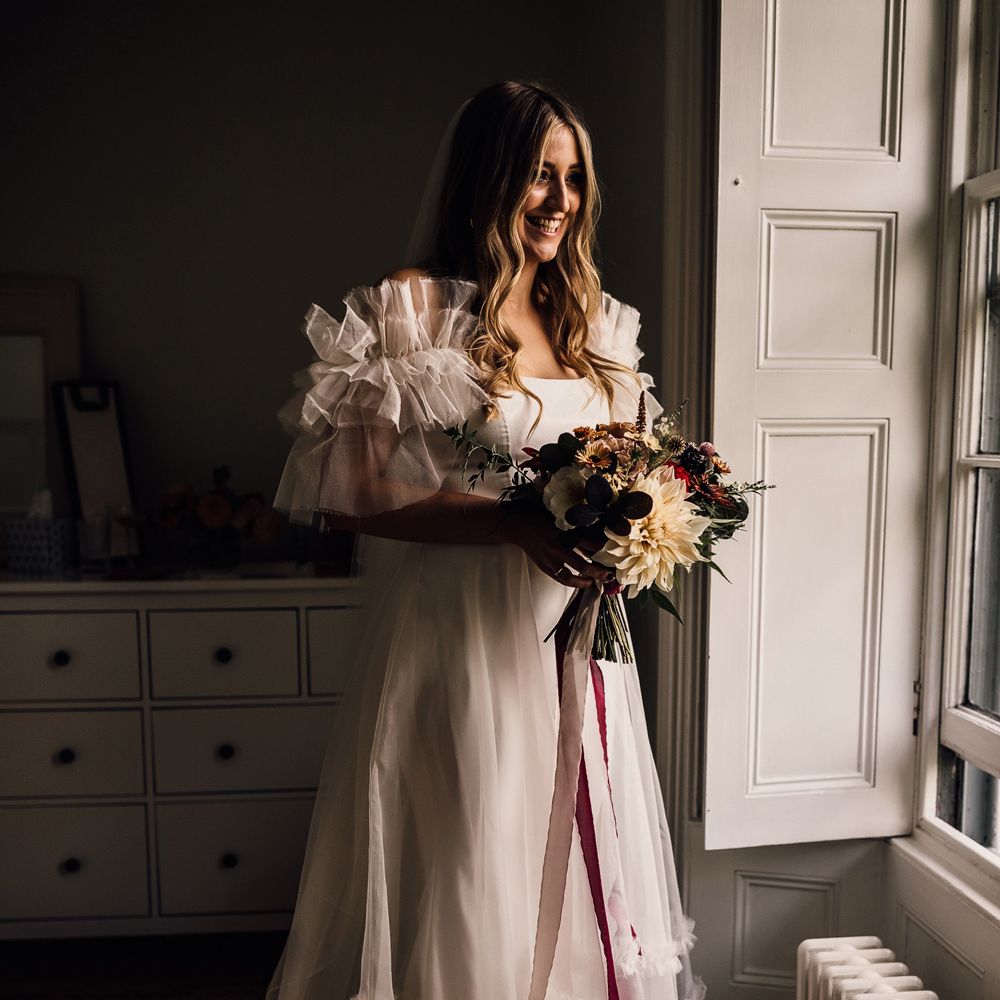 Bride in white Halfpenny London Mayfair dress with ruffle sleeves holding floral bouquet stands by window before church wedding ceremony in Cornwall