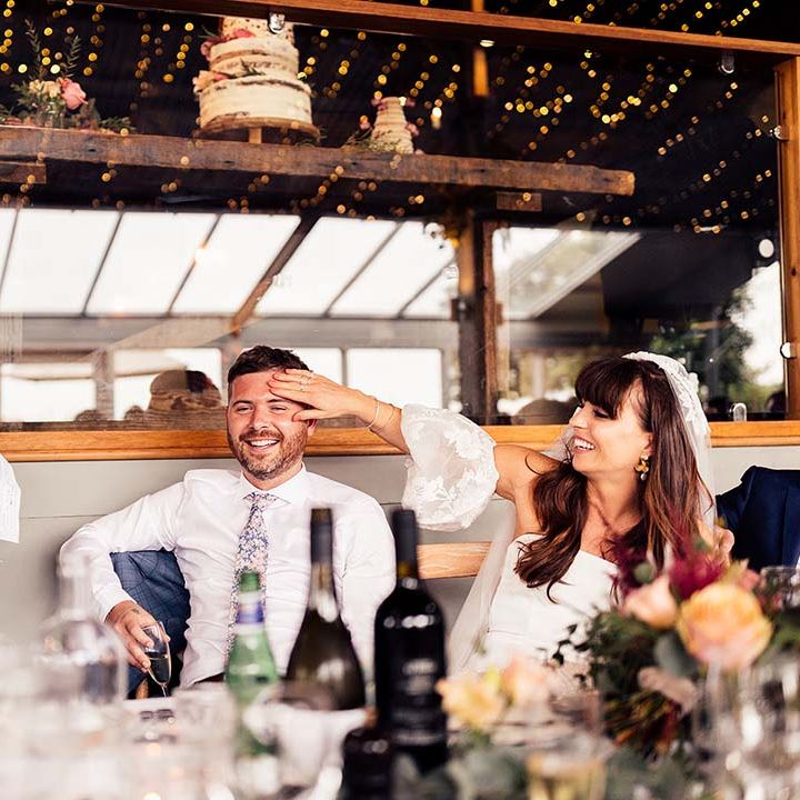 Bride in a strapless wedding dress with lace detachable sleeves feeling her grooms head as the best man delivers his wedding speech 
