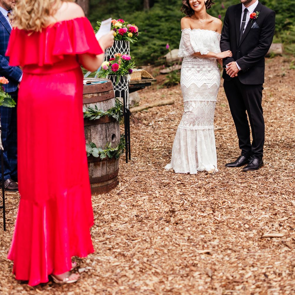 Bride in strapless Grace Loves Lace wedding dress and flower Cornwall stands with groom in black suit and bridesmaid in pink bridesmaid dress performs wedding reading during outdoor ceremony