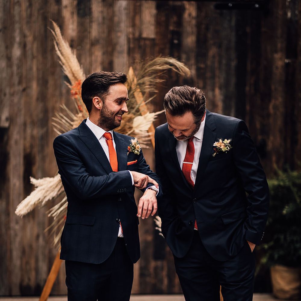 Groom in a navy suit with brown bridges and an orange tie standing at the wooden triangle altar decorated with pampas grass with his best man 