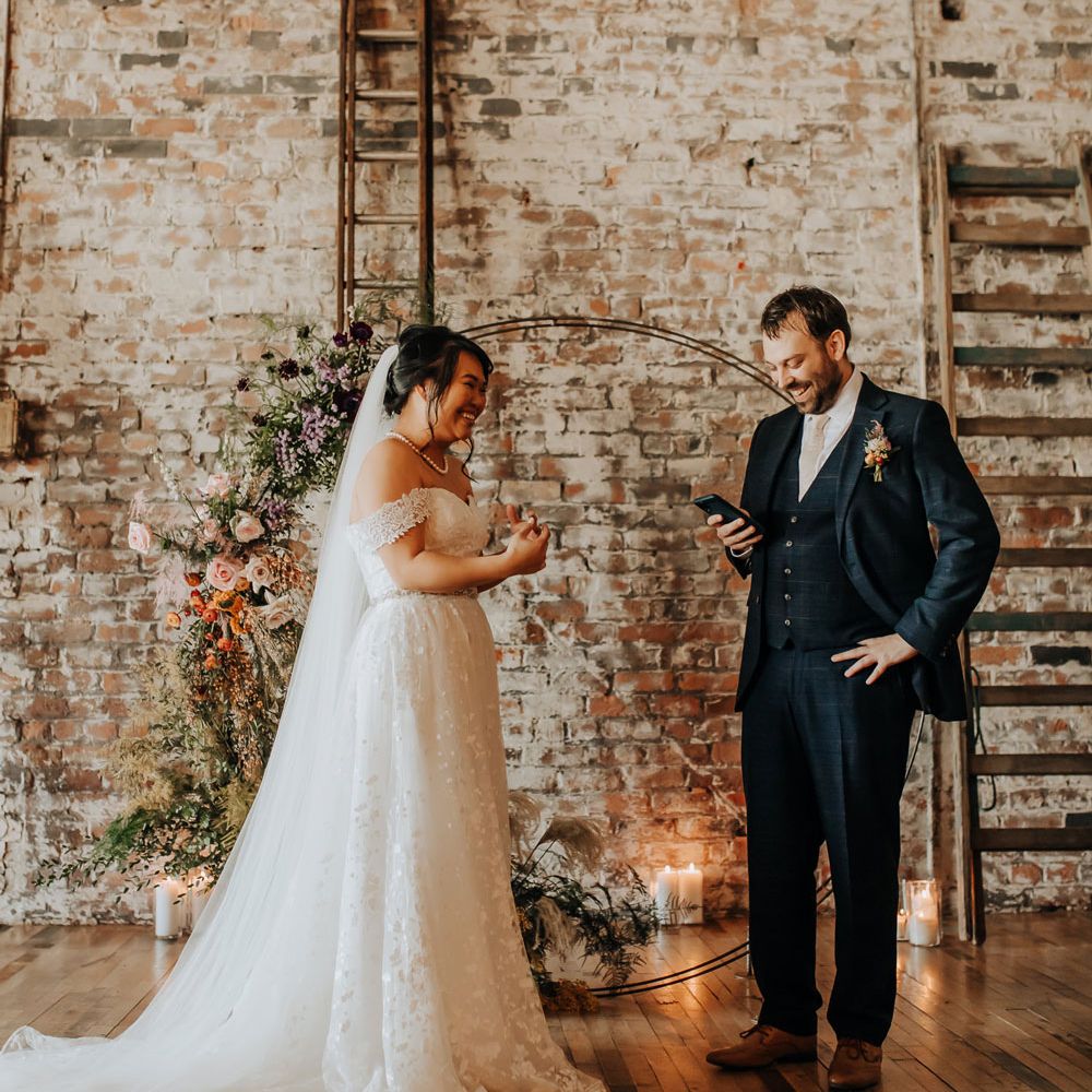 Bride in bardot wedding dress and veil stands by floral moongate whilst groom in blue suit reads vows off his phone at wedding ceremony in Liverpool