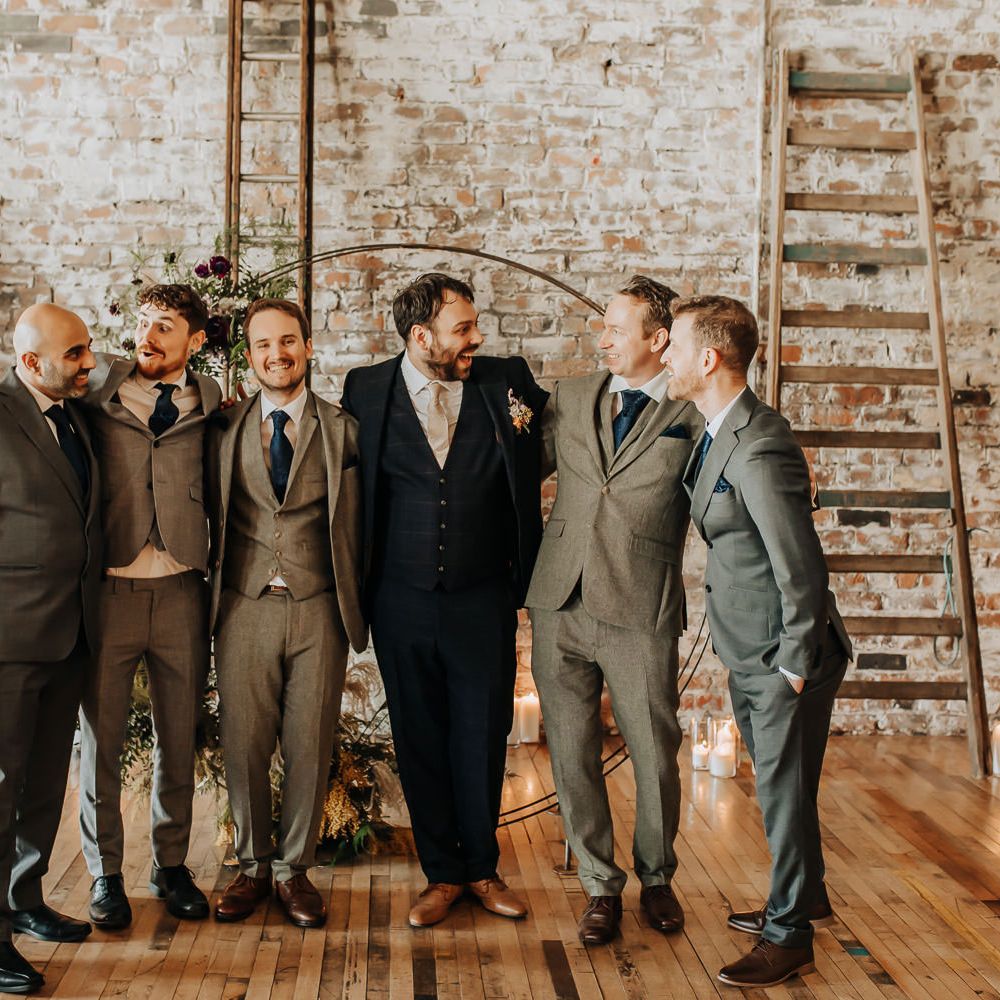 Groom in blue check three piece suit stands with groomsmen in grey three piece suits and blue ties in front of floral moongate
