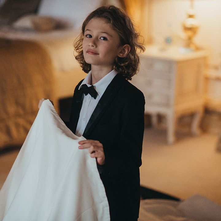 Page boy wears black tie and holds train of Suzanne Neville wedding gown