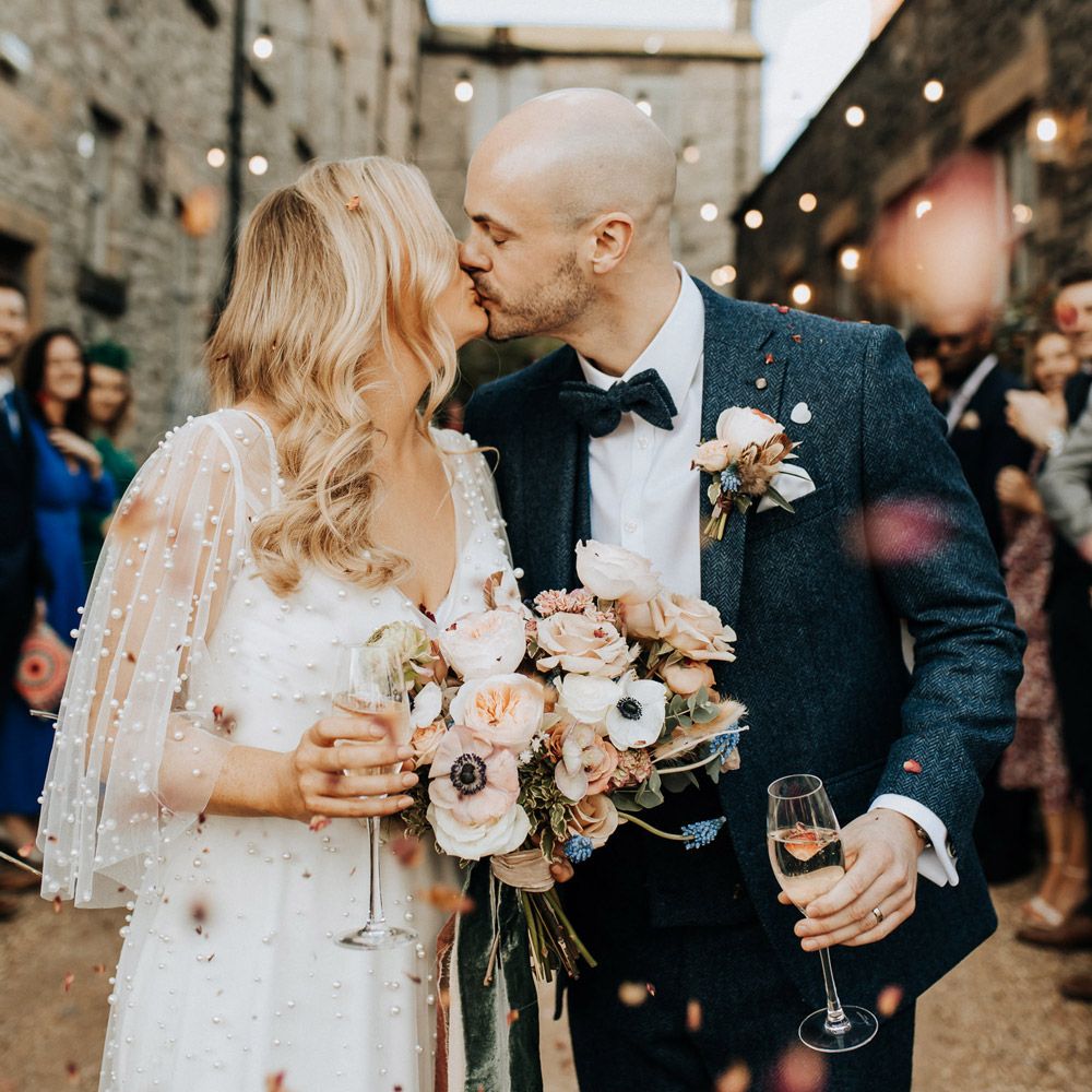 Bride with blonde hair and wedding dress with short sleeves encrusted with pearls holding pastel wedding bouquet kisses groom in navy tweed wedding suit with buttonhole