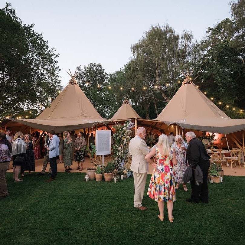 Wedding guests mingle outside of the beige Buffalo Tipi for garden wedding in Bath with fairy lights