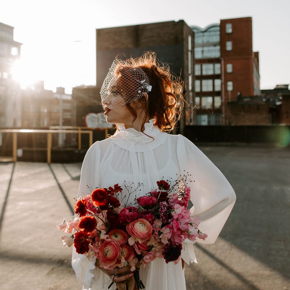 Bride in a high neck wedding dress with long sleeves wearing a birdcage veil with pearls on holding a romantic red bouquet during golden hour 