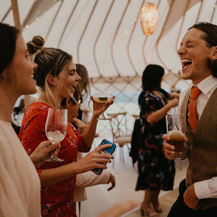 Groomsman in brown waistcoat and orange tie holding a pint laughs with wedding guests