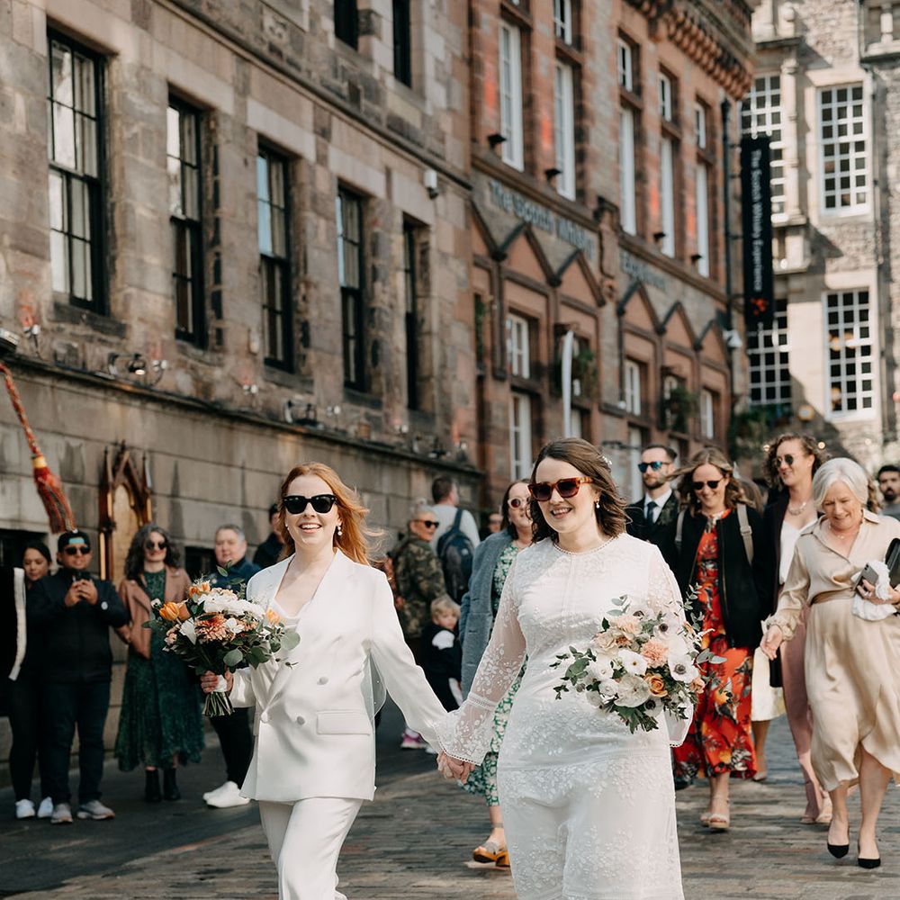 Brides wearing sunglasses walk with one another on their wedding day holding hands and carrying floral bouquets 