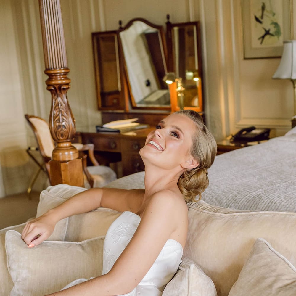 Bride with updo hairstyle lounges back on the sofa in her Jesus Peiro wedding dress with brocade pattern