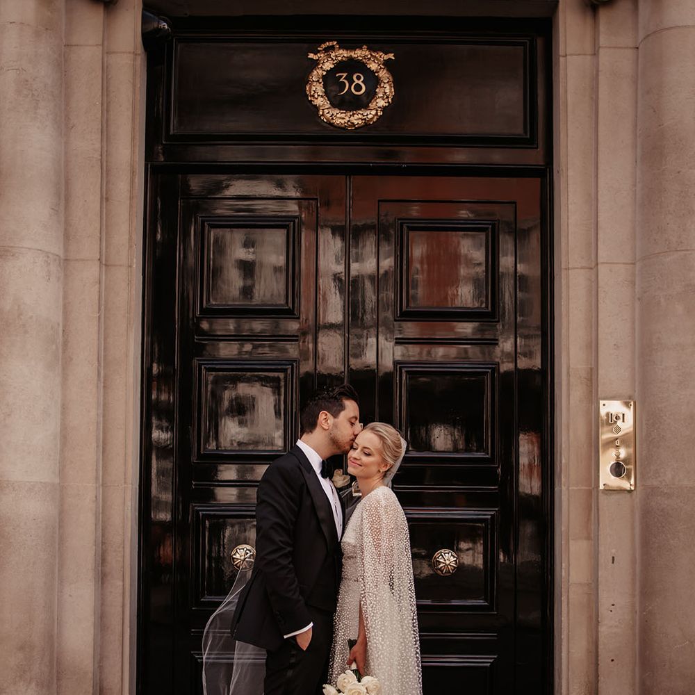Groom in black tie kisses the bride on the forehead with bride in sparkly Jesus Peiro cape 