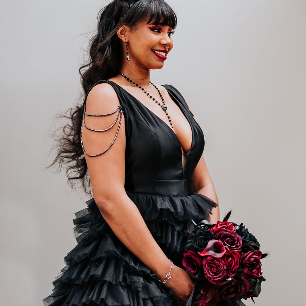 Black bride wears black ruffled wedding dress complete with chain detailing to shoulders and wears her hair in space buns with skull bridal crown