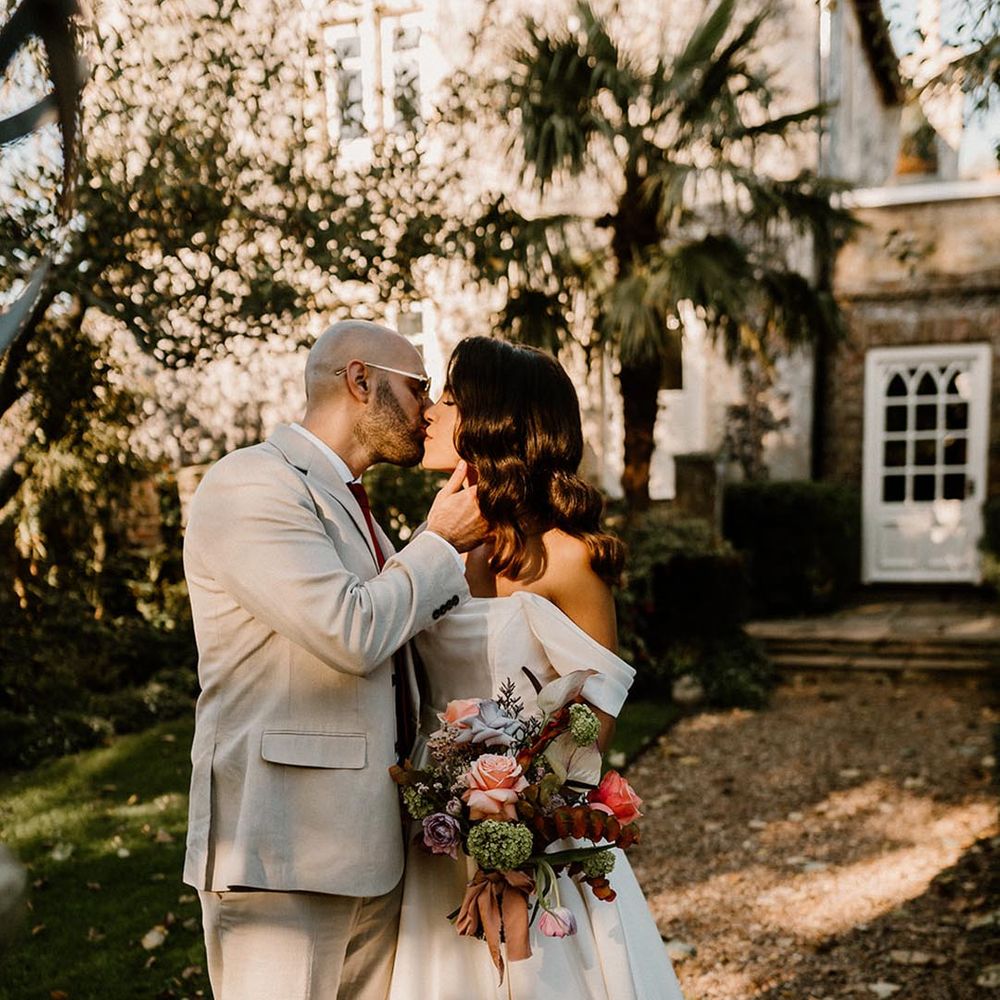 Groom in a pale grey su8it and dark red tie kisses the bride in an off the shoulder wedding dress and Hollywood waves