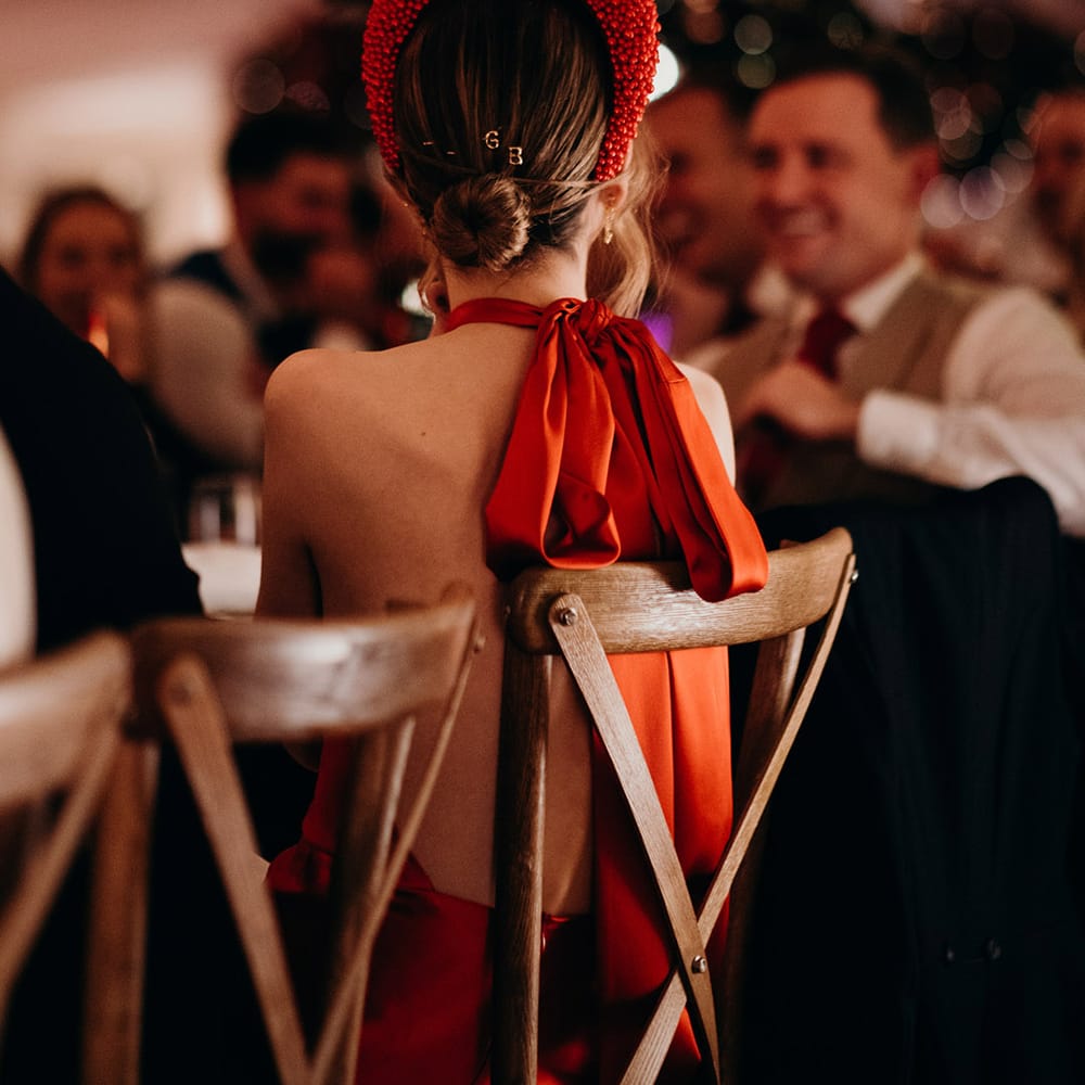 Bride sitting at reception wearing red silk wedding dress with cape, large red embellished head band, letter hair clips and large silk bow