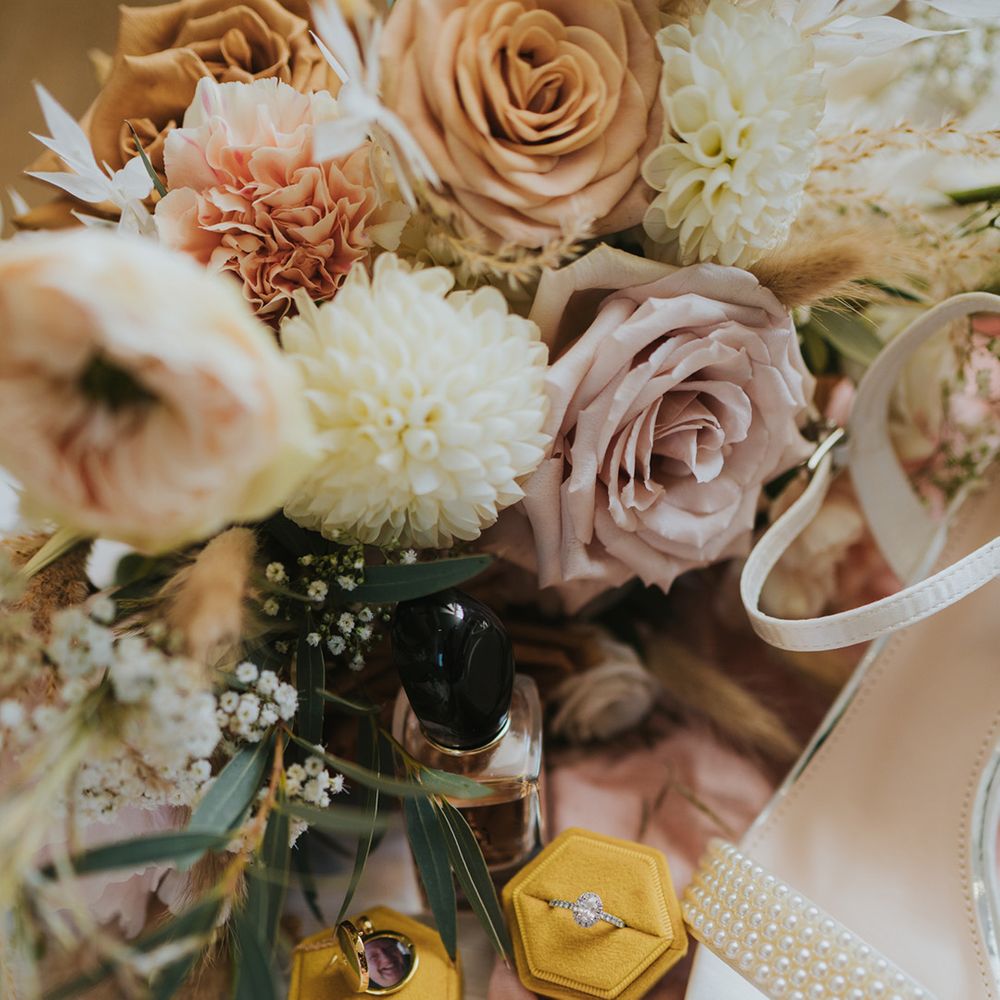 Diamond wedding ring in yellow box beside pearl embellished bridal shoes and floral bouquet