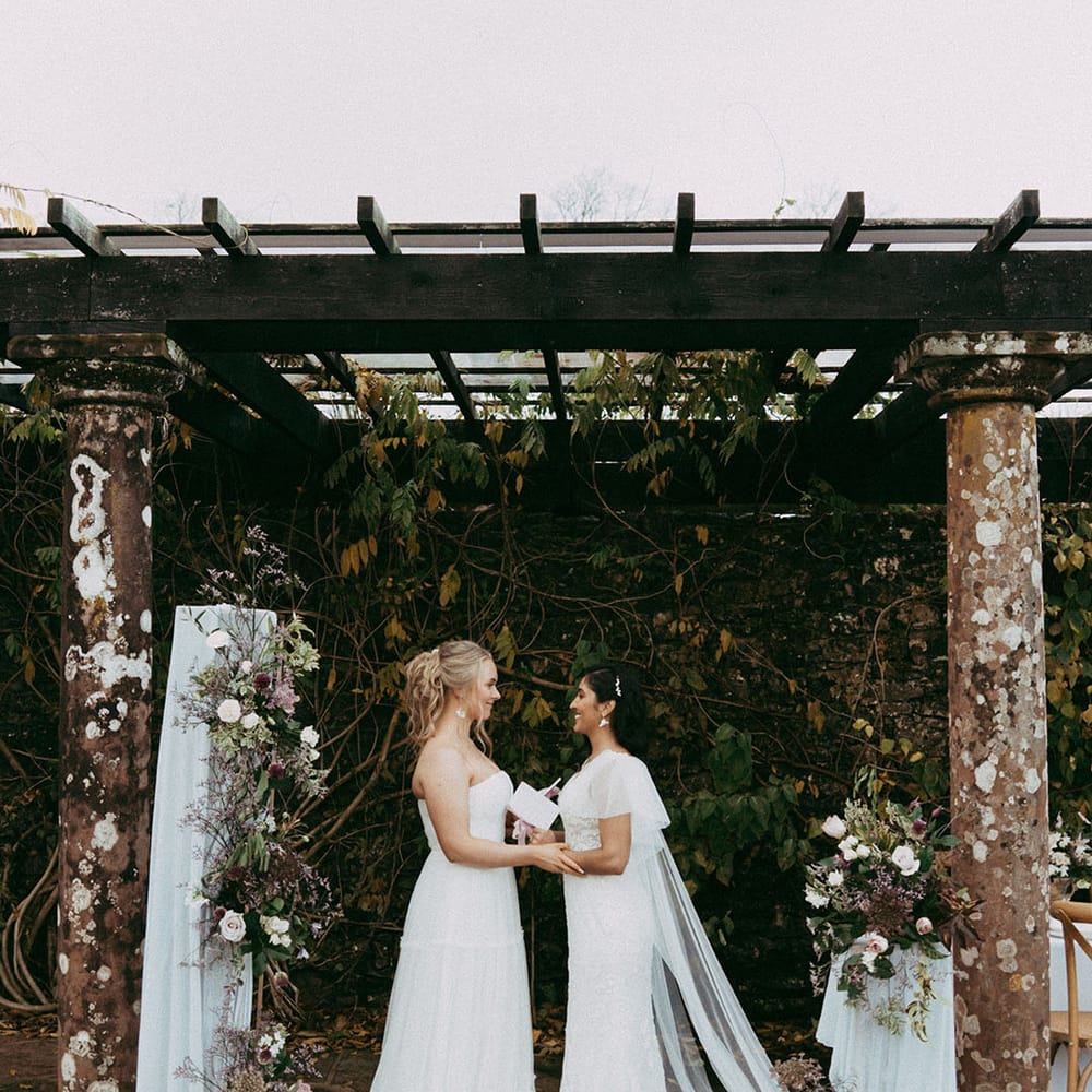 Brides in sleeveless lace wedding dress and off the shoulder long sleeve wedding dress holding hands at the alter with mixed floral decorations with white carnations, purple tulips, roses and foliage 