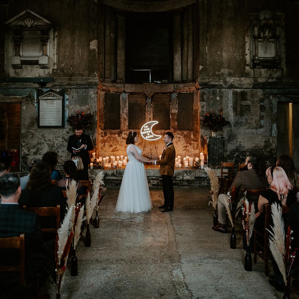 Bride and Groom stand at alter with moon neon sign in the centre at industrial wedding venue in London