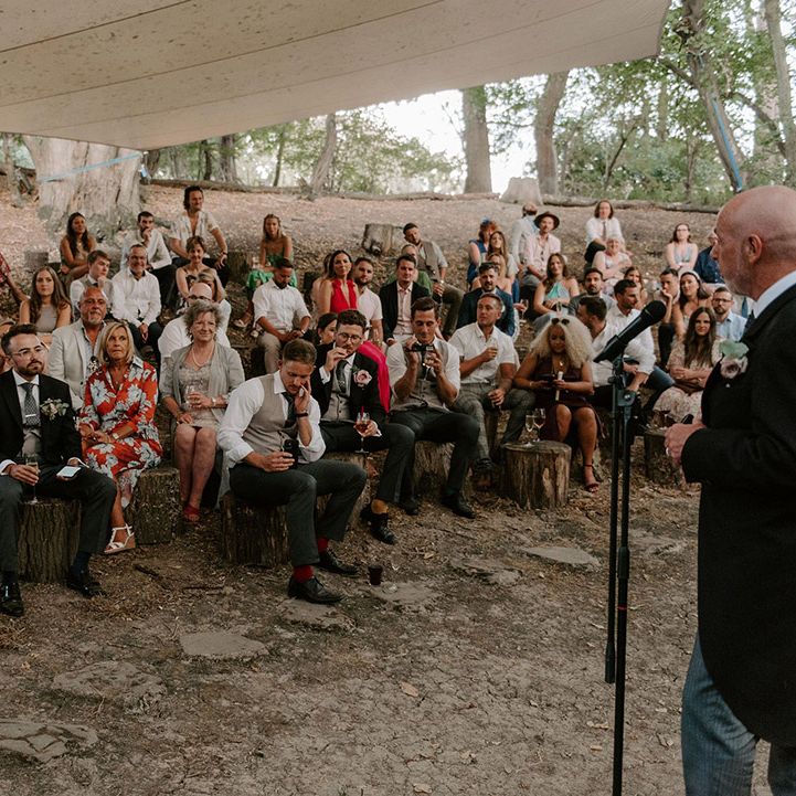 Groomsman reads out a wedding speech as the bride and groom and wedding guests sit and listen in secret outdoor area 