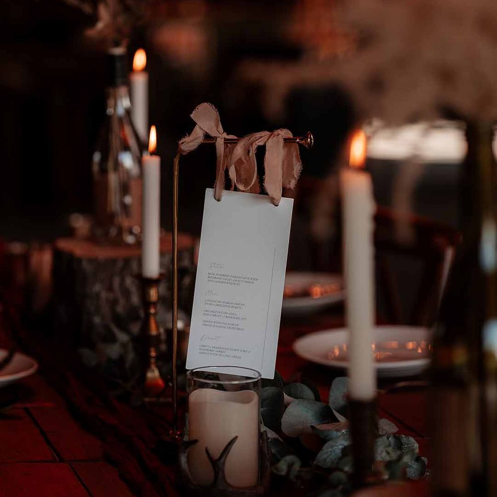 Autumnal boho wedding tablescape with white tapered candles in gold candlesticks, foliage table runner decor, pillar candles in glass candle jars, and minimalistic wedding menu on golden stand with neutral peach ribbon