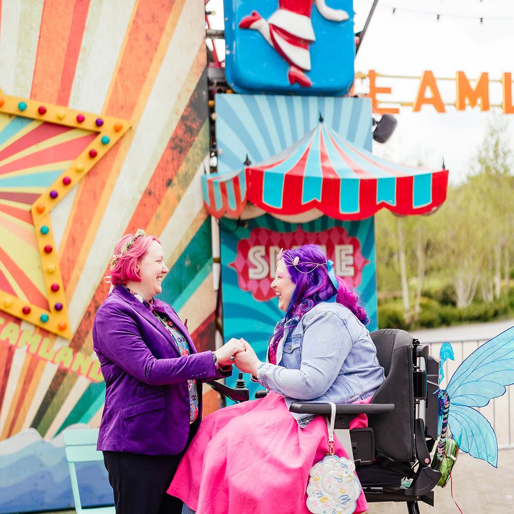 Bride in wheelchair with sparkly blue fairy wings attached wearing personalised denim jacket, pink wedding dress and blue tights holding hands with bride in  purple velvet blazer and black suit trousers with purple bowtie, pink patterned pocket square and colourful patterned shirt 