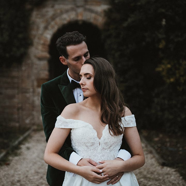 Bride in off the shoulder lace wedding dress being embraced from behind by the groom 