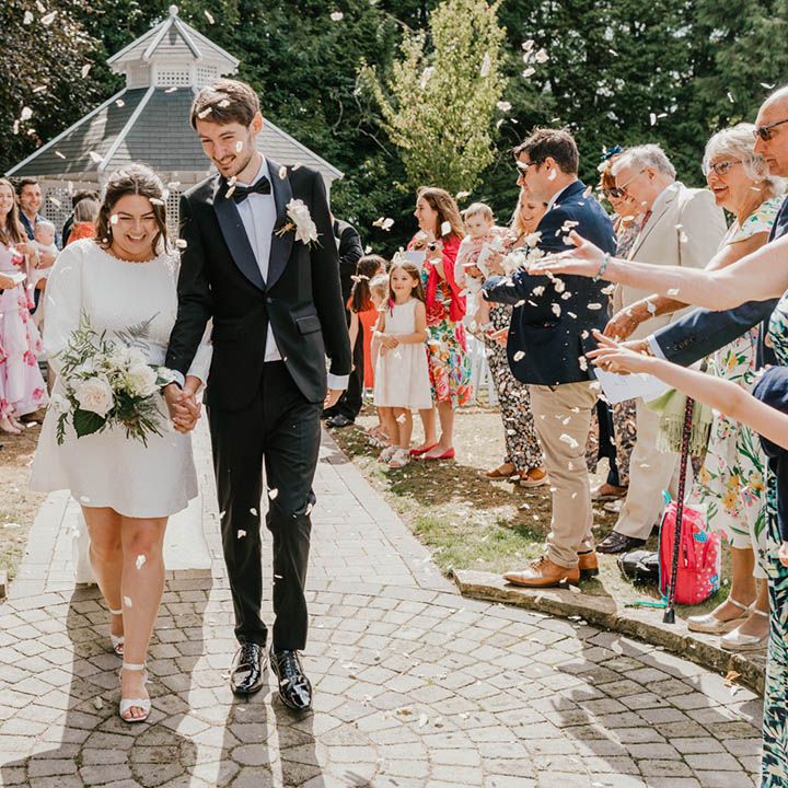 The bride in a short wedding dress with long sleeves and a train with bow detail walks back down the aisle with the groom after their outdoor wedding ceremony at Hayne House