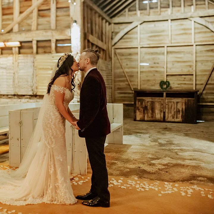 The bride in an elegant lace wedding gown kisses the groom in a deep red velvet suit jacket for their first look moment at Milling Barn