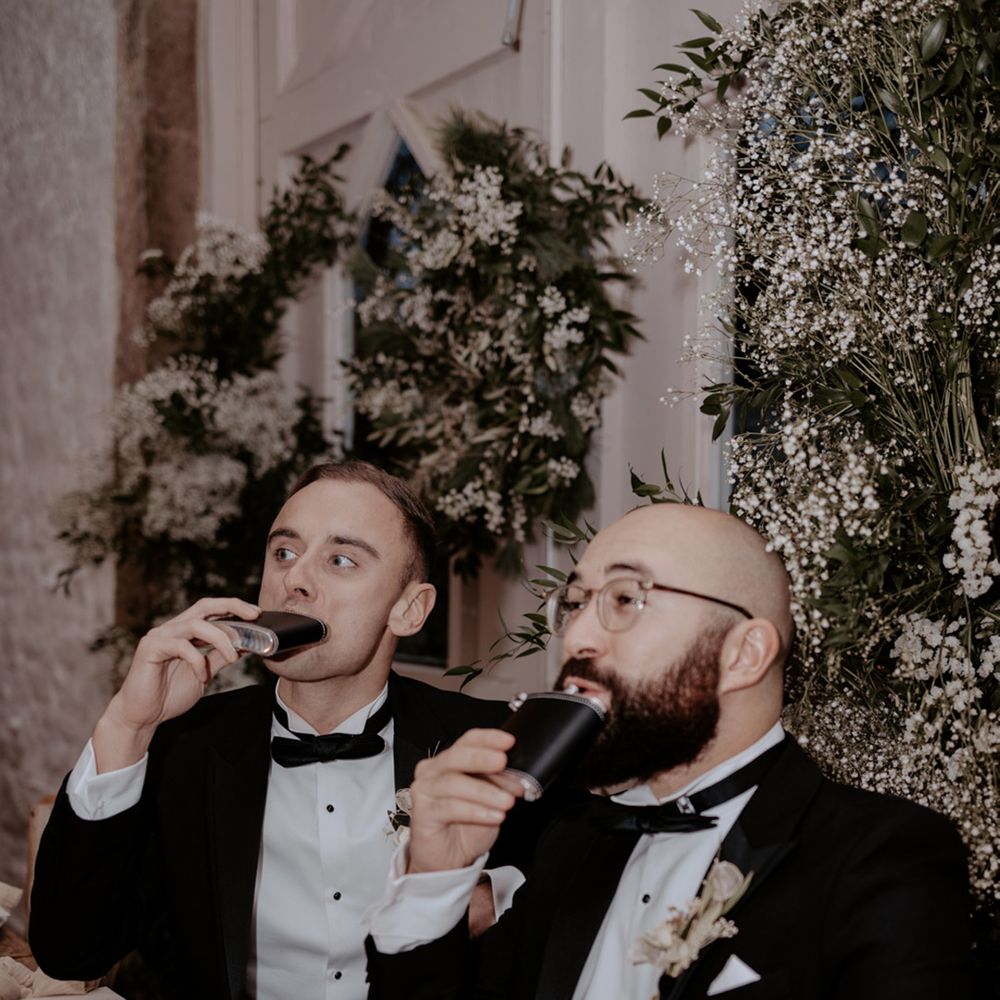Groomsmen in black tuxedos drink alcohol from their matching flasks 