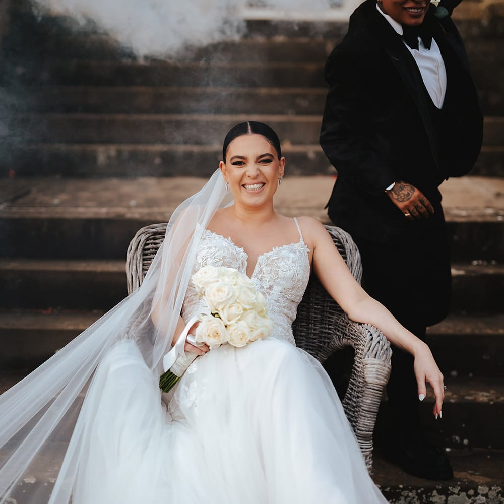 Bride in lace sleeveless v neck Enzoani wedding dress with tulle finish and church length veil sitting on wicker chair during smoke bomb photoshoot at Grittleton House wedding venue 