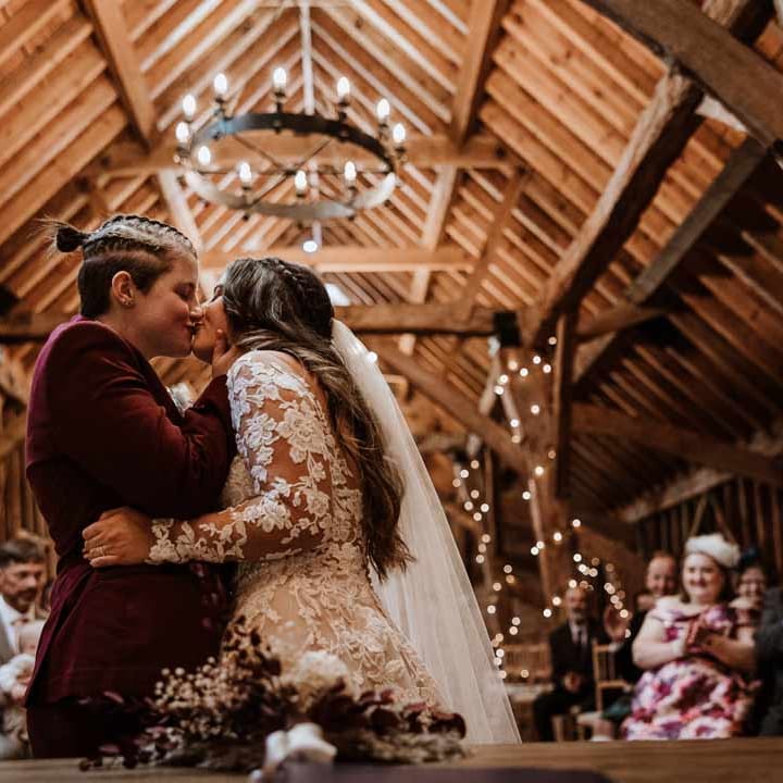 Bride in lace illusion sleeve wedding dress with overlay doing first kiss with bride in maroon bridal suit at LGBTQ+ wedding at Bassmead Manor Barns wedding venue 