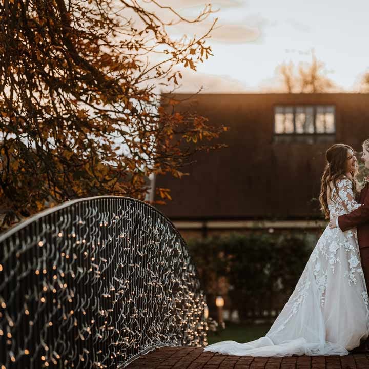 Bride in long sleeve lace illusion sleeve wedding dress with overlay standing on a bridge at Bassmead Manor Barns with bride in maroon bridal suit with grey waistcoat, dusky pink tie and autumnal dried flower boutonniere 