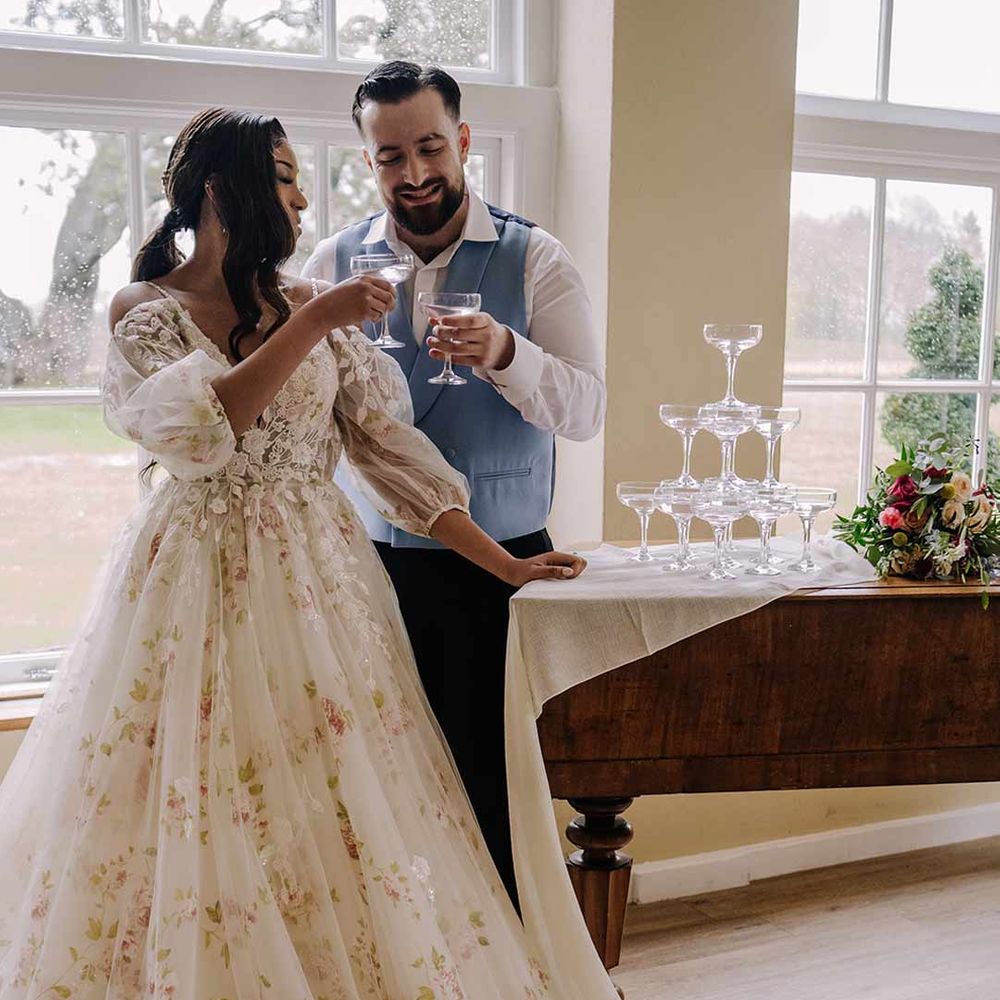 Bride in v-neck long sleeve shoulder cut out 3d appliqué wedding dress with flecks of gold having a glass of champagne with groom in cornflower blue waistcoat and black suit trousers at Braxted Park wedding venue 