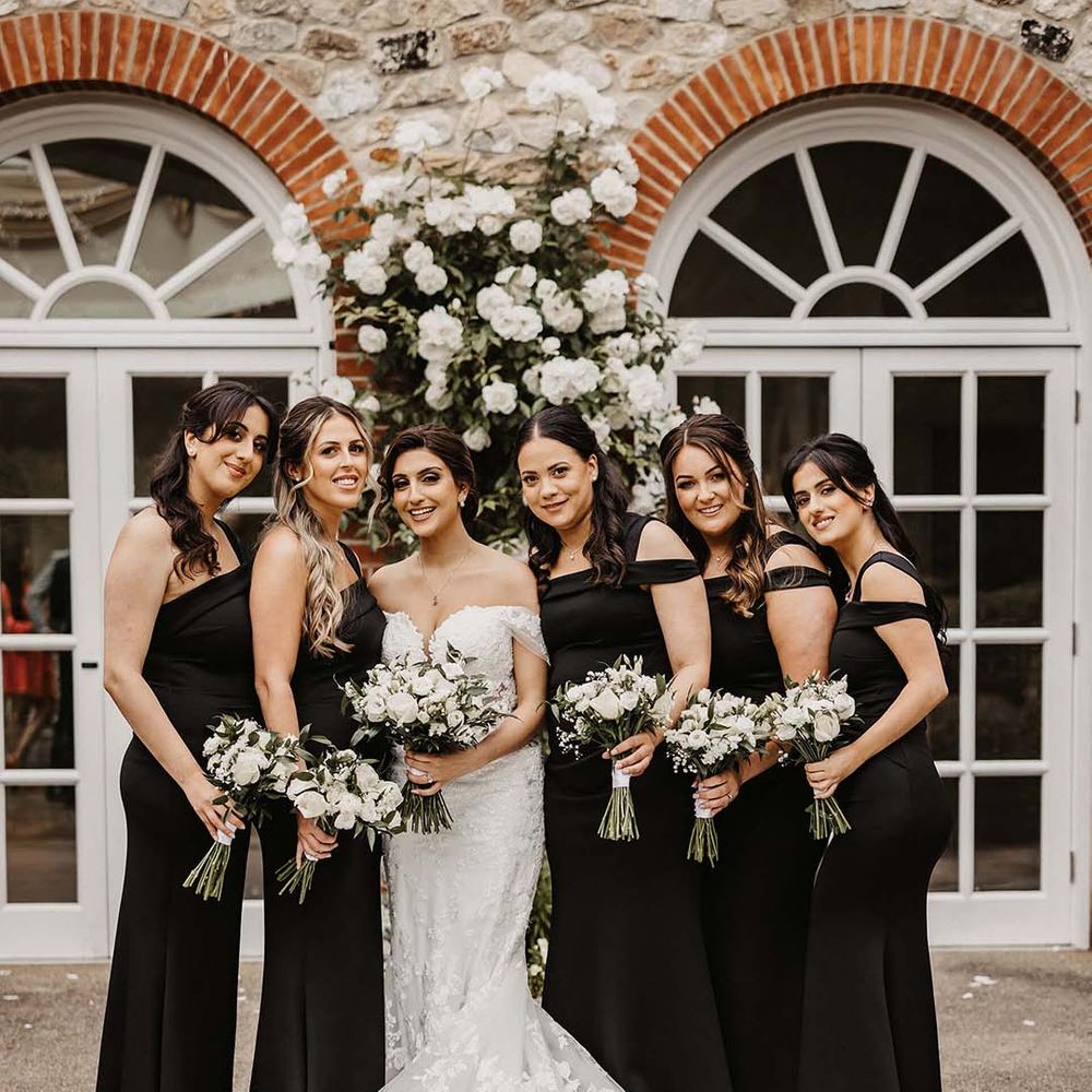 Bride in off the shoulder lace wedding dress with puddle train standing with bridesmaids in black strappy bridesmaid dresses 