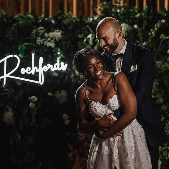 Bride in sweetheart neckline lace wedding dress standing and laughing with groom in classic black three piece grooms suit with bow tie and white rose and foliage boutonniere in church in front of foliage wall and large neon wedding sign 