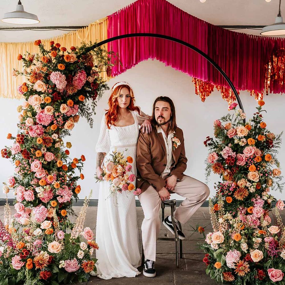 Bride in three quarter length puff sheer sleeve wedding dress with delicate polka dot overlay and square neck and white 3D applique polka dot headband posing with groom in brown blazer, white shirt, grey suit trousers and vans shoes underneath circular floral arch with vibrant flowers and foliage attached and pink, red, yellow and metallic wedding streamers behind