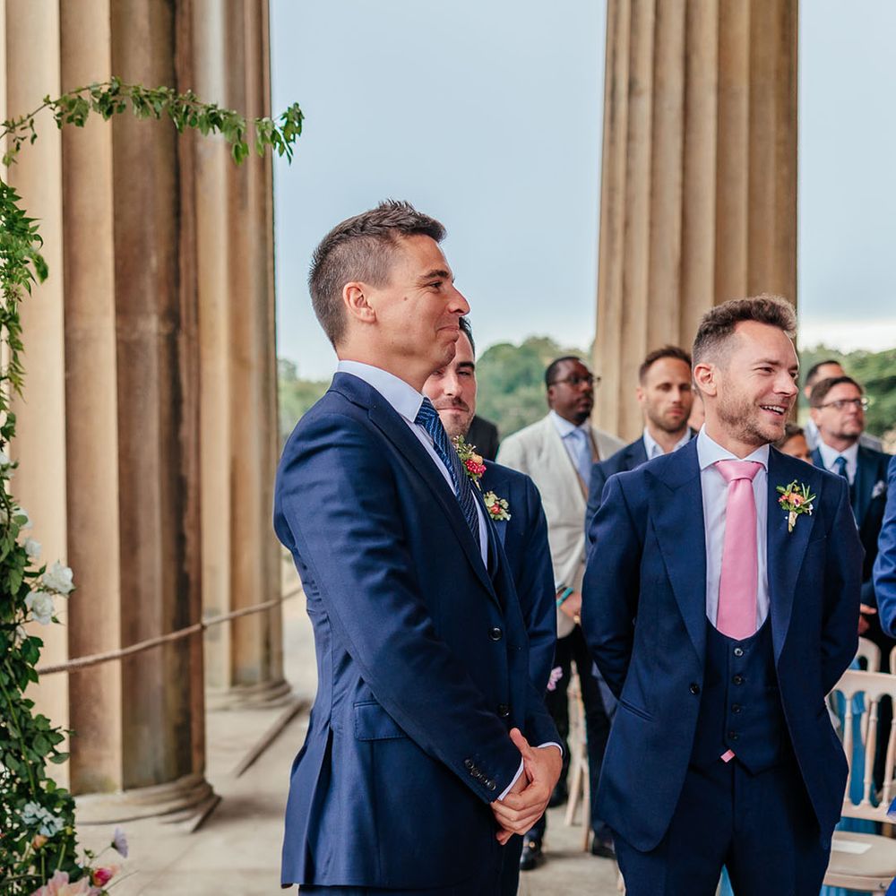Groom in blue suit smiles as he sees the bride walking down the aisle 