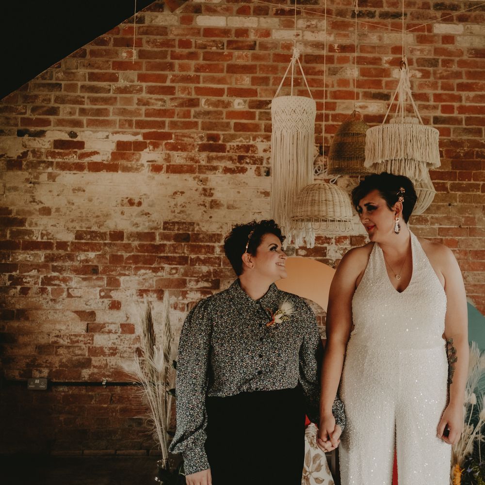 Bride in a jumpsuit and bride in a patterned shirt and trousers holding hands 