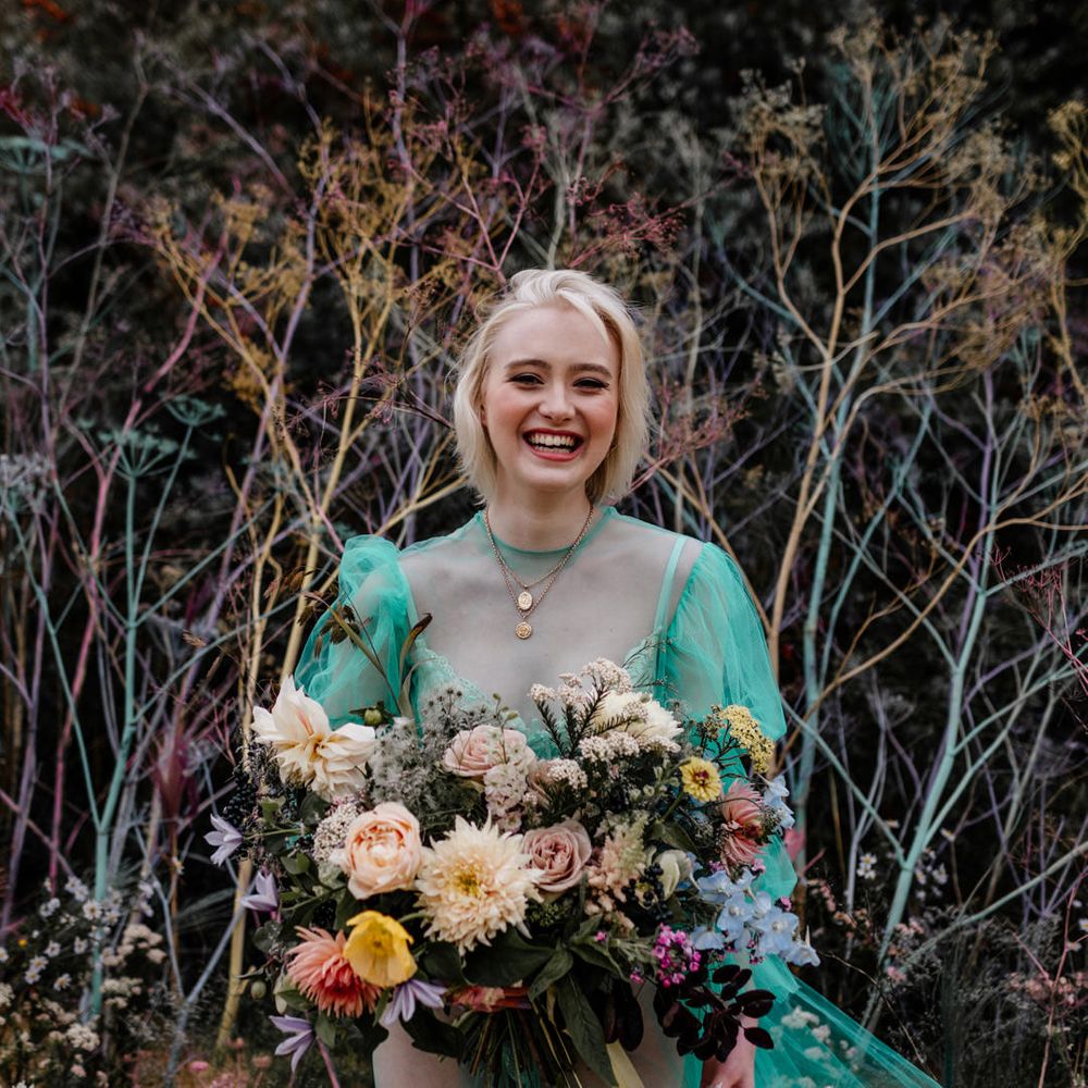 Smiling blonde bride in a high neck sheer wedding dress wearing gold chains and holding a colourful wedding bouquet 