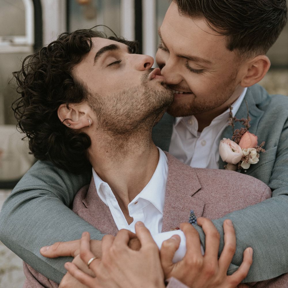 Grooms kiss outdoors whilst holding arms around one another
