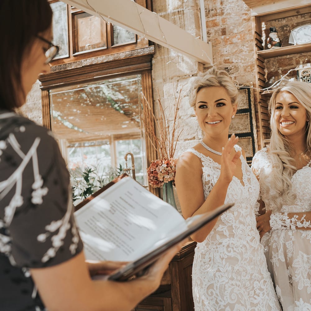 Two brides in white dresses with blonde hair kiss as a guest performs a reading at their wedding at Crab and Lobster Fairytale Wedding.
