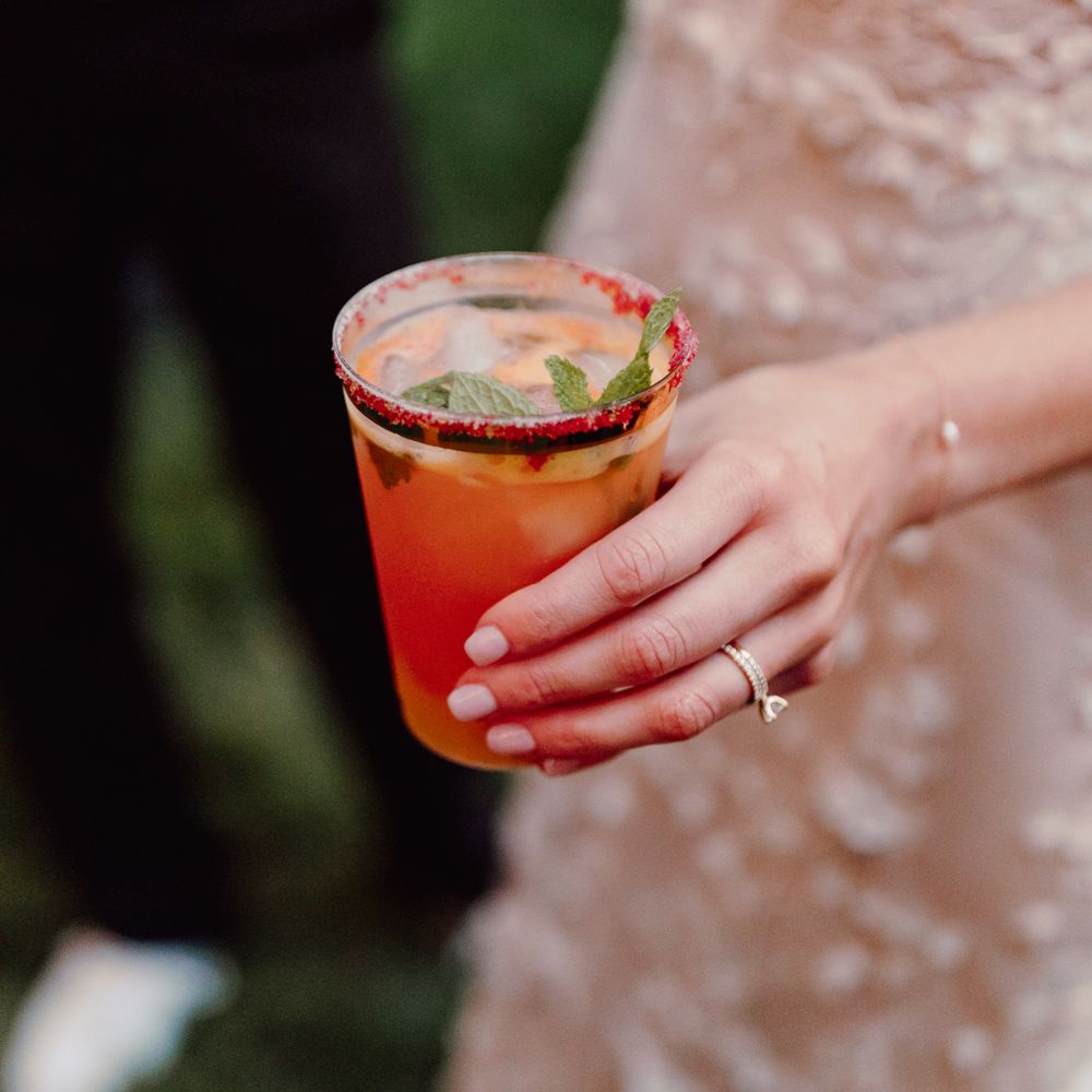 Bride in white embroidered Made With Love wedding dress holds tumbler filled with ice, orange drink and mint at backyard wedding