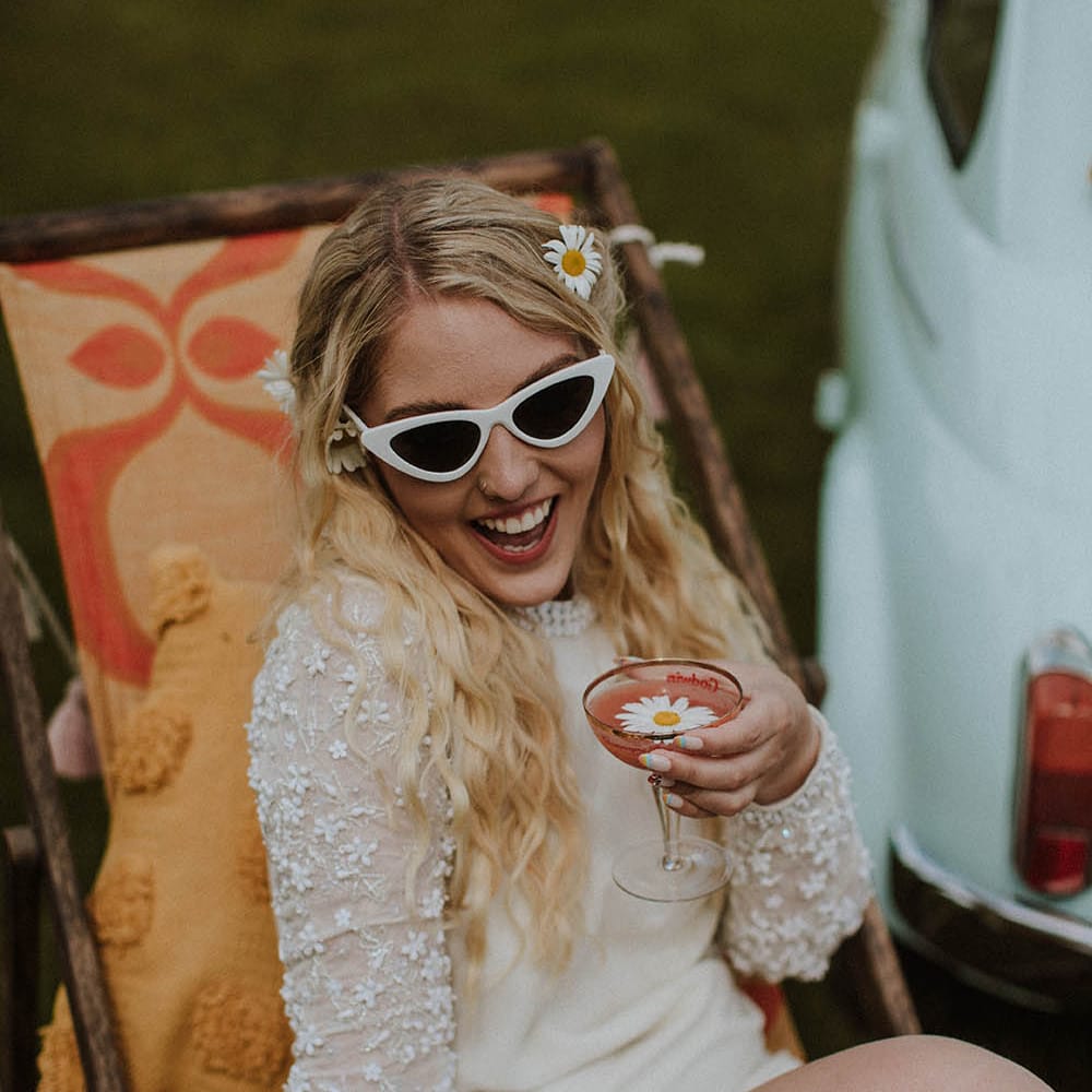 Bride in a short wedding dress with appliqué long sleeves holding a coupe glass and wearing white winged sunglasses 