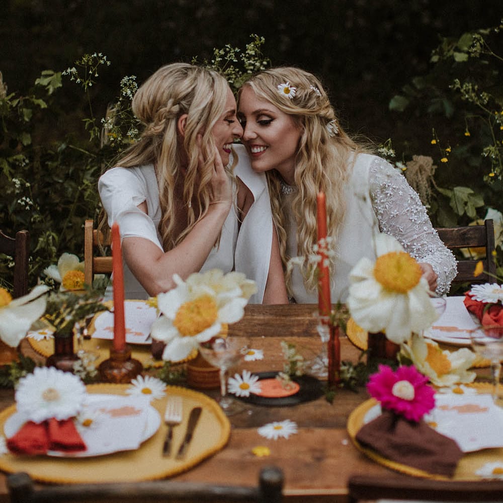 two boho brides with long wavy hair sitting at a sixties decorated wedding reception tablescape